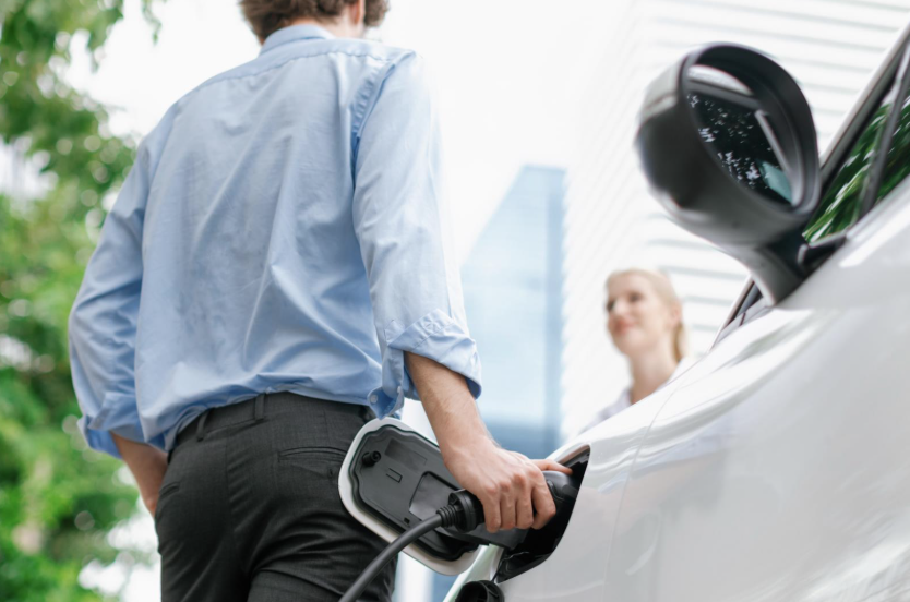Focus hand insert EV charger to electric vehicle at public charging point in car park with blur business people in backdrop, eco-friendly lifestyle by rechargeable car for progressive concept. 