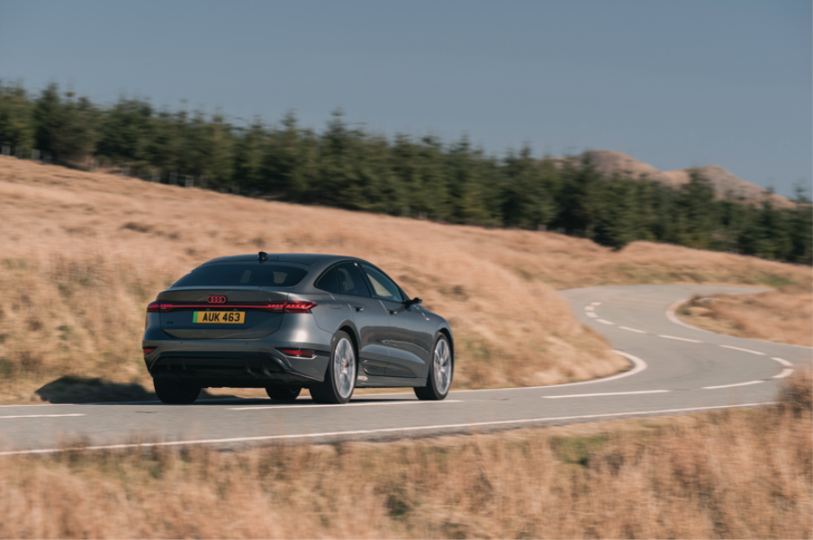 Grey Audi Q6 e-tron driving along a winding rural road through dry hills.