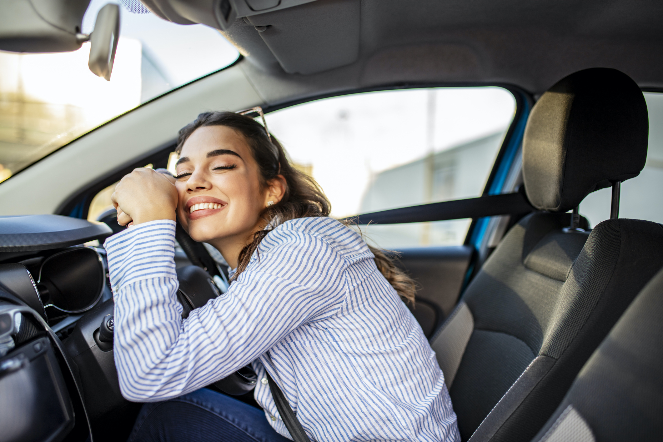 Young Woman Embracing Her New Car. Excited young woman and her new car indoors. Young and cheerful woman enjoying new car hugging steering wheel sitting inside.  